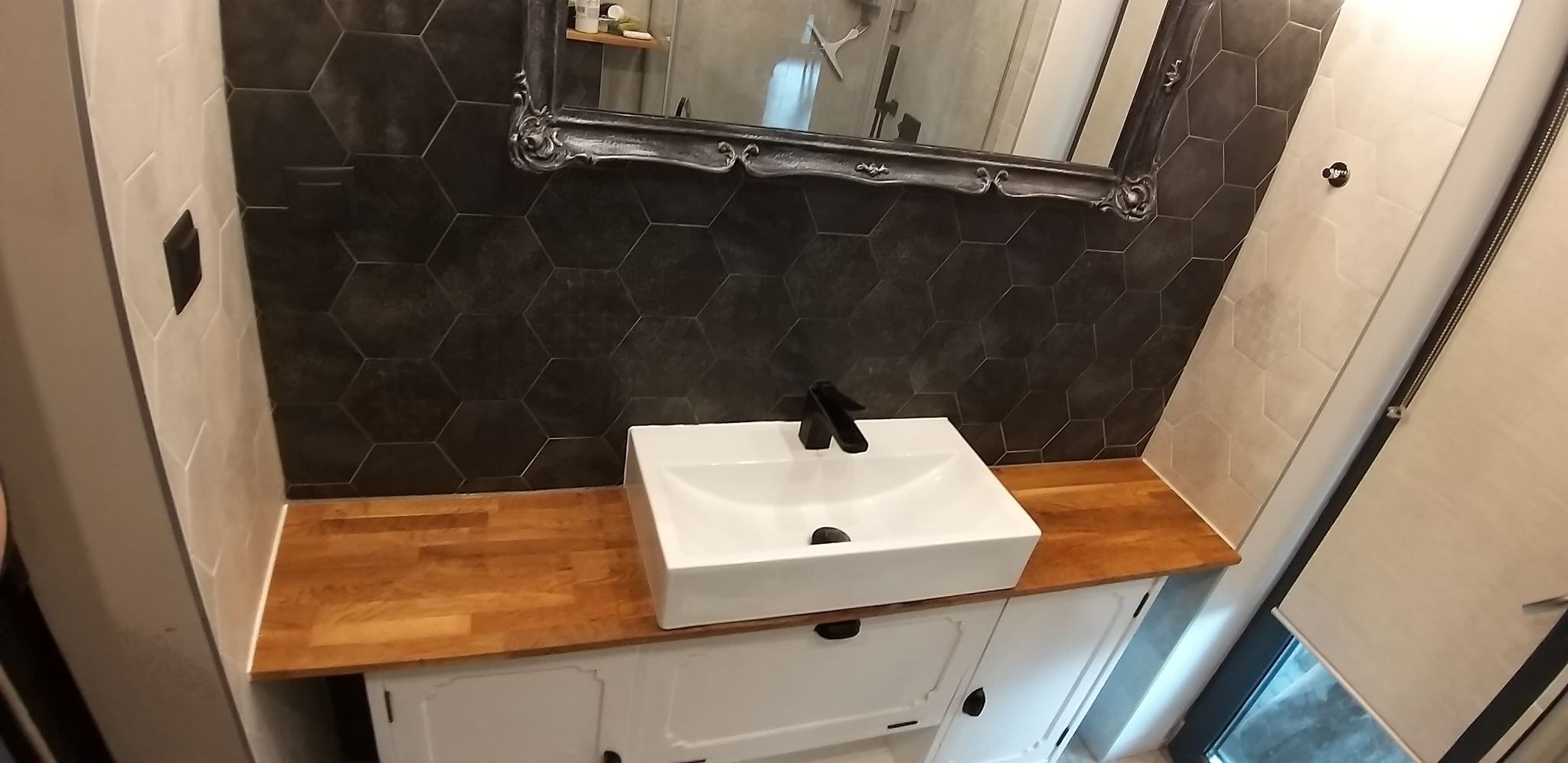 Umywalka nablatowa z czarną armaturą na drewnianym blacie w łazience z ciemnymi, heksagonalnymi płytkami na ścianie. W tle widoczne lustro w ozdobnej ramie.