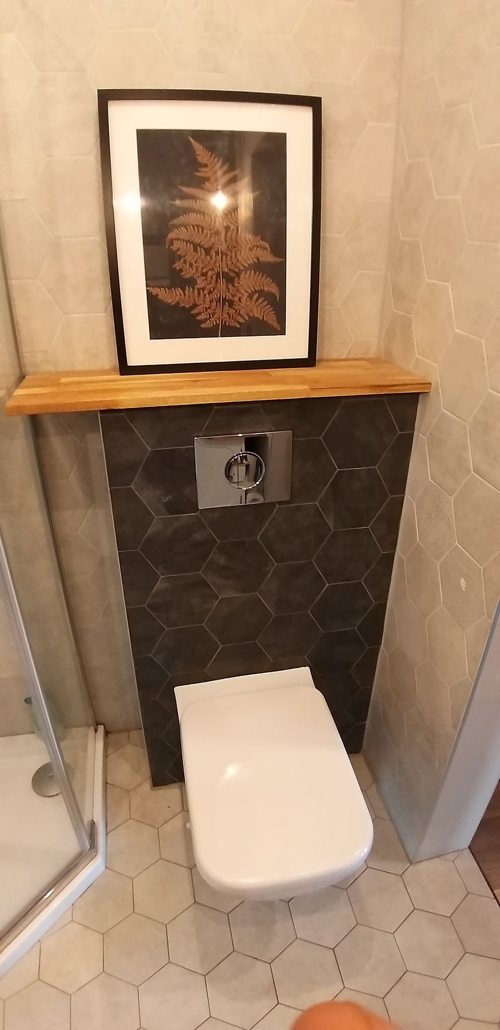 Nowoczesna łazienka z zabudowanym stelażem WC wykończonym ciemnymi płytkami hexagonalnymi, dębowy blat nad stelażem i wisząca grafika z motywem paproci. Na podłodze jasne płytki hexagonalne...