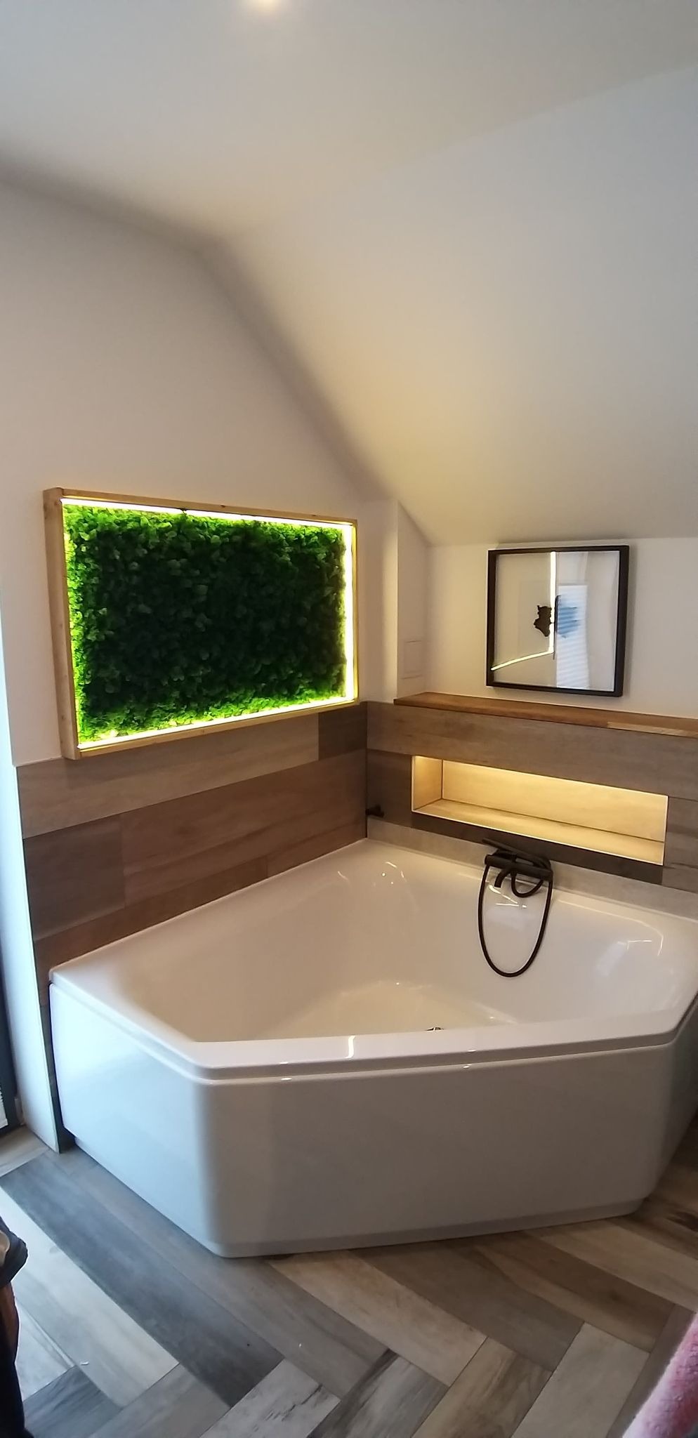 Nowoczesna łazienka w skandynawskim stylu z narożną wanną, drewnopodobnymi płytkami na ścianach i podłodze, podświetlaną półką ścienną oraz panelem z chrobotka norweskiego z oświetleniem LED.