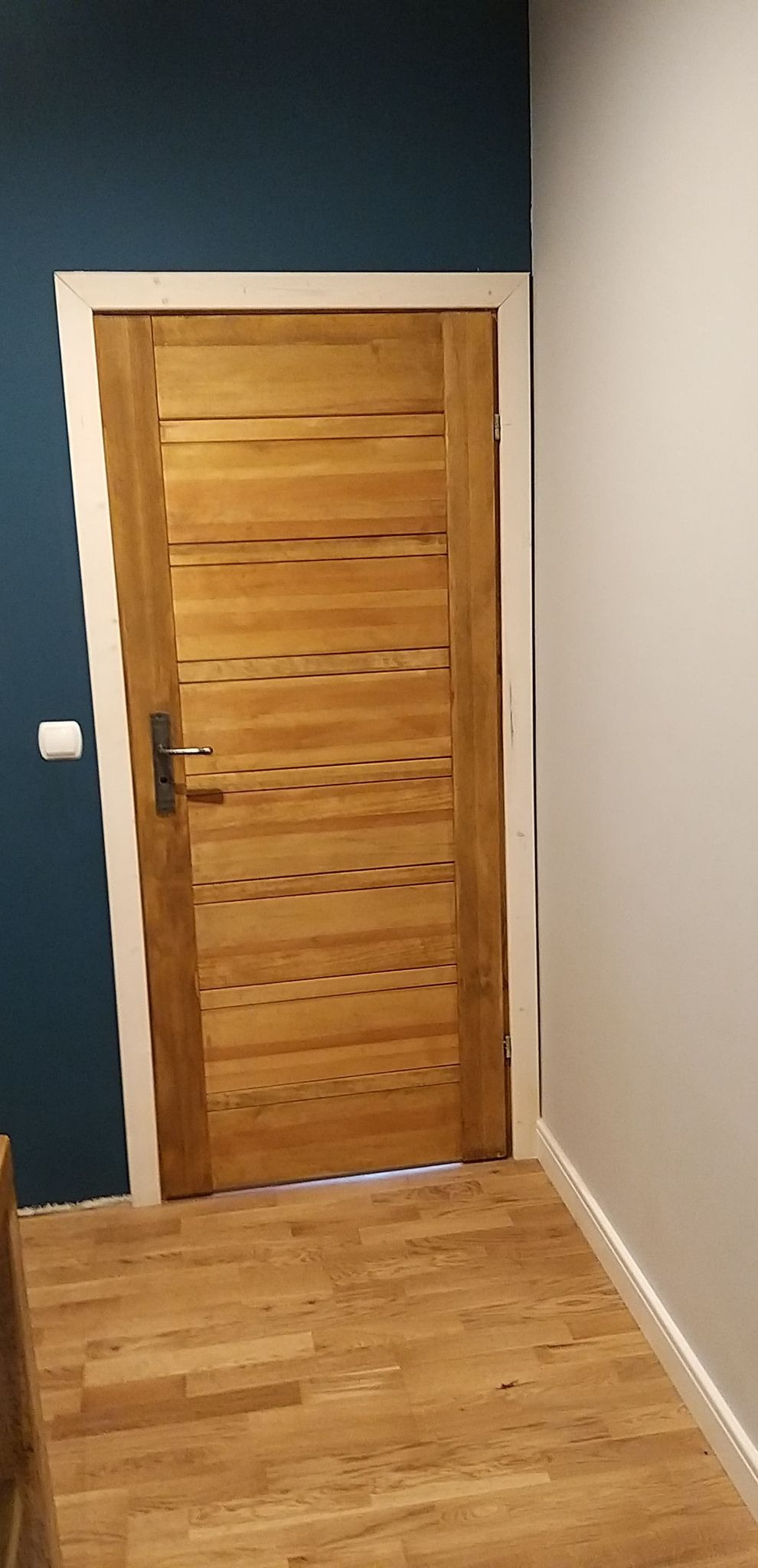 Zamknięte drewniane drzwi z poziomymi panelami w białej ościeżnicy, zamontowane na tle ściany w kolorze granatowym i białym, z widoczną drewnianą podłogą i białą listwą przypodłogową.