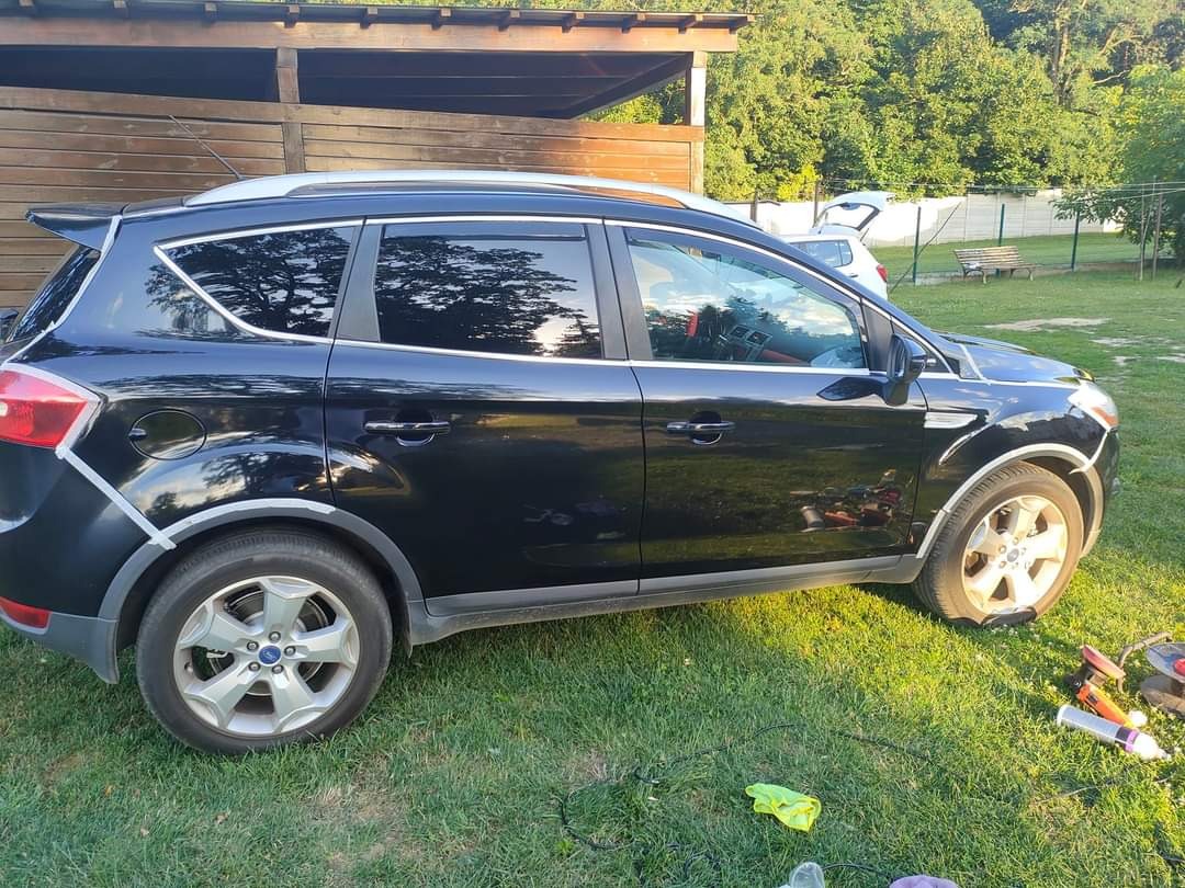 Czarny samochód Ford Kuga z taśmą maskującą wokół nadkoli, przygotowywany do polerowania na trawniku, widoczne narzędzia i preparaty.