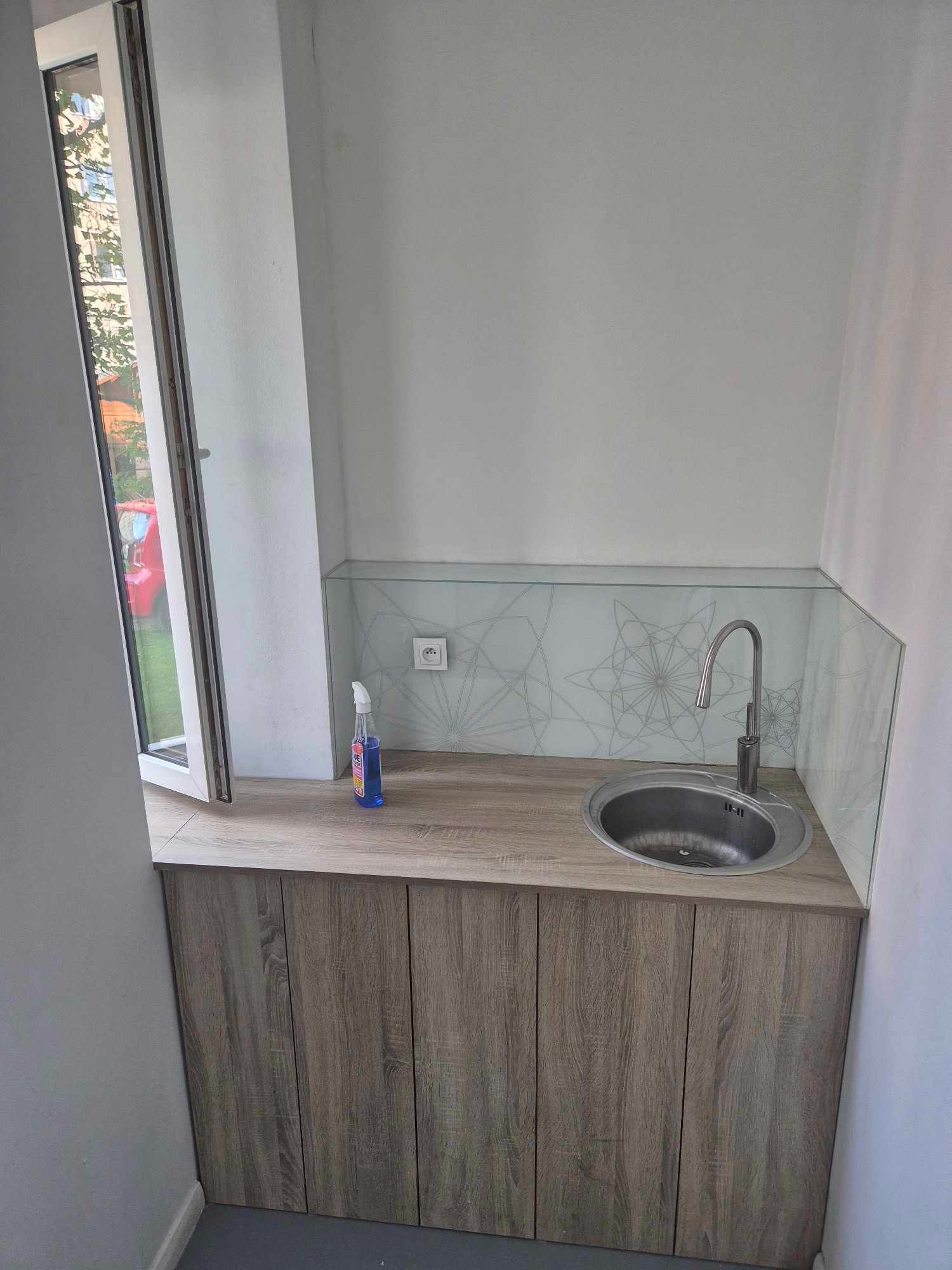 Kompaktowa umywalka ze stali nierdzewnej z wysoką baterią, osadzona w blacie z drewnopodobnego laminatu, z otwartym oknem po lewej stronie i butelką niebieskiego płynu do czyszczenia obok, na tle...