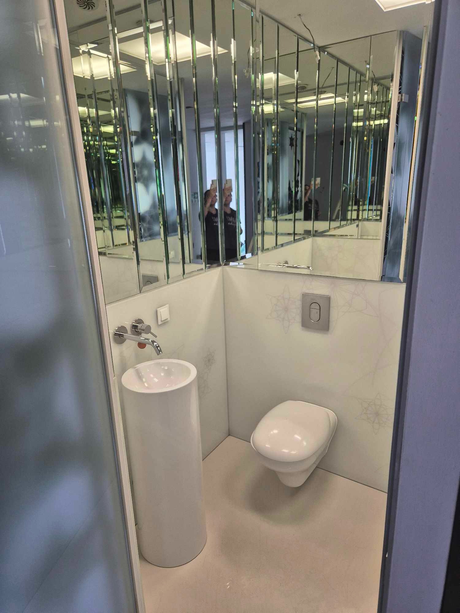 Wnętrze małej, nowoczesnej toalety z umywalką na postumencie i podwieszaną miską WC, wykończonej lustrami i delikatnym wzorem na ścianach.