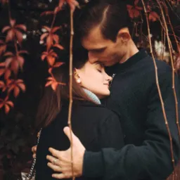 Zakochana para w jesiennej scenerii: mężczyzna całuje kobietę w czoło, otoczeni bordowymi liśćmi winobluszczu.