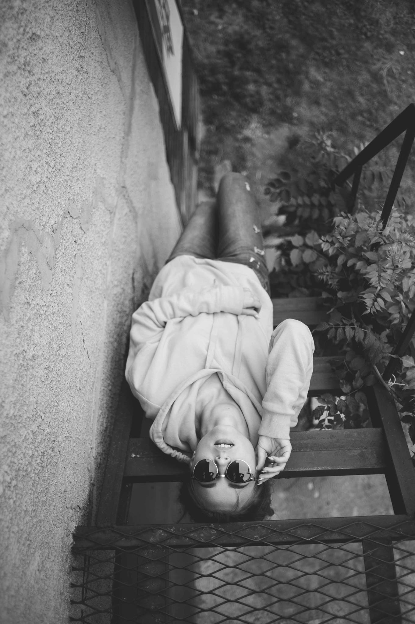 Czarno-białe zdjęcie kobiety w okularach przeciwsłonecznych, leżącej na metalowych schodach w bluzie z kapturem, z bliska widoczna struktura betonowej ściany i roślinność w tle.