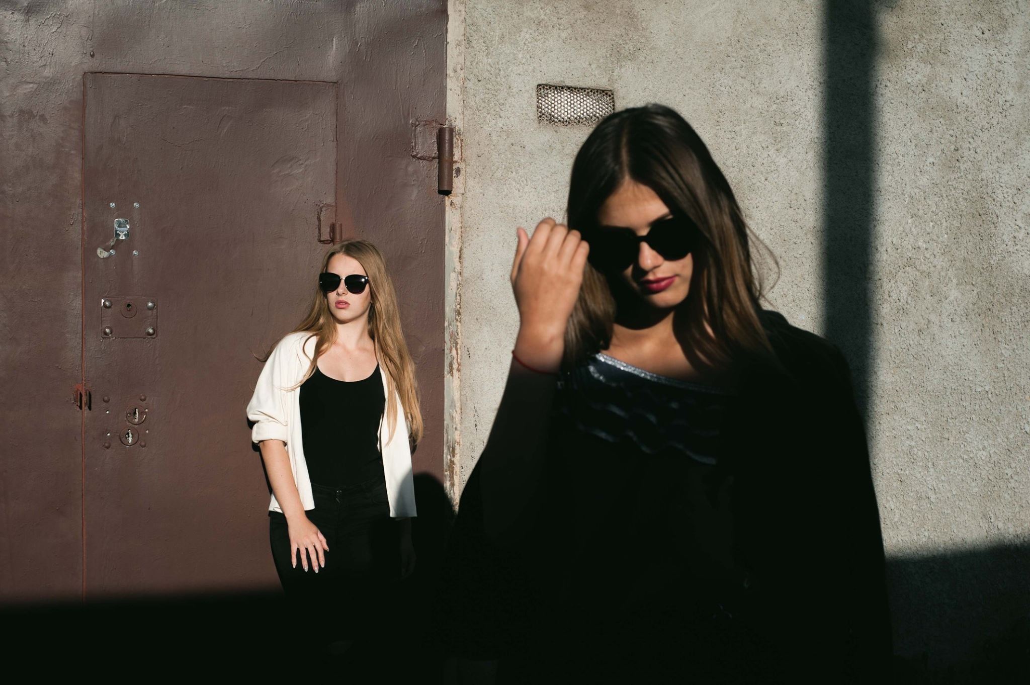 Dwie młode kobiety w okularach przeciwsłonecznych pozują na tle surowych ścian; jedna oparta o brązowe metalowe drzwi, druga w cieniu z ręką we włosach.