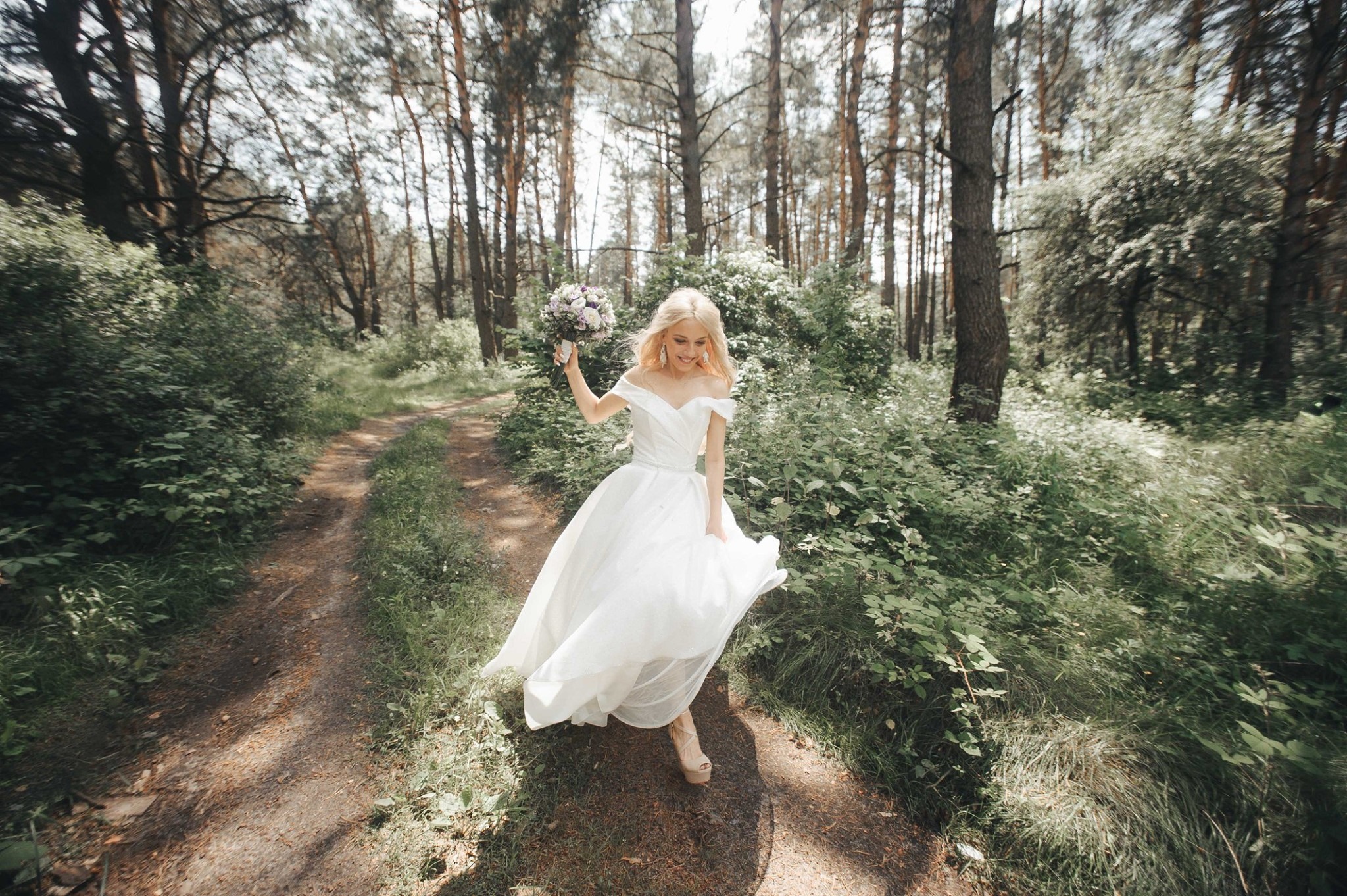 Uśmiechnięta panna młoda w białej sukni, biegnąca ścieżką w lesie, trzyma bukiet kwiatów.