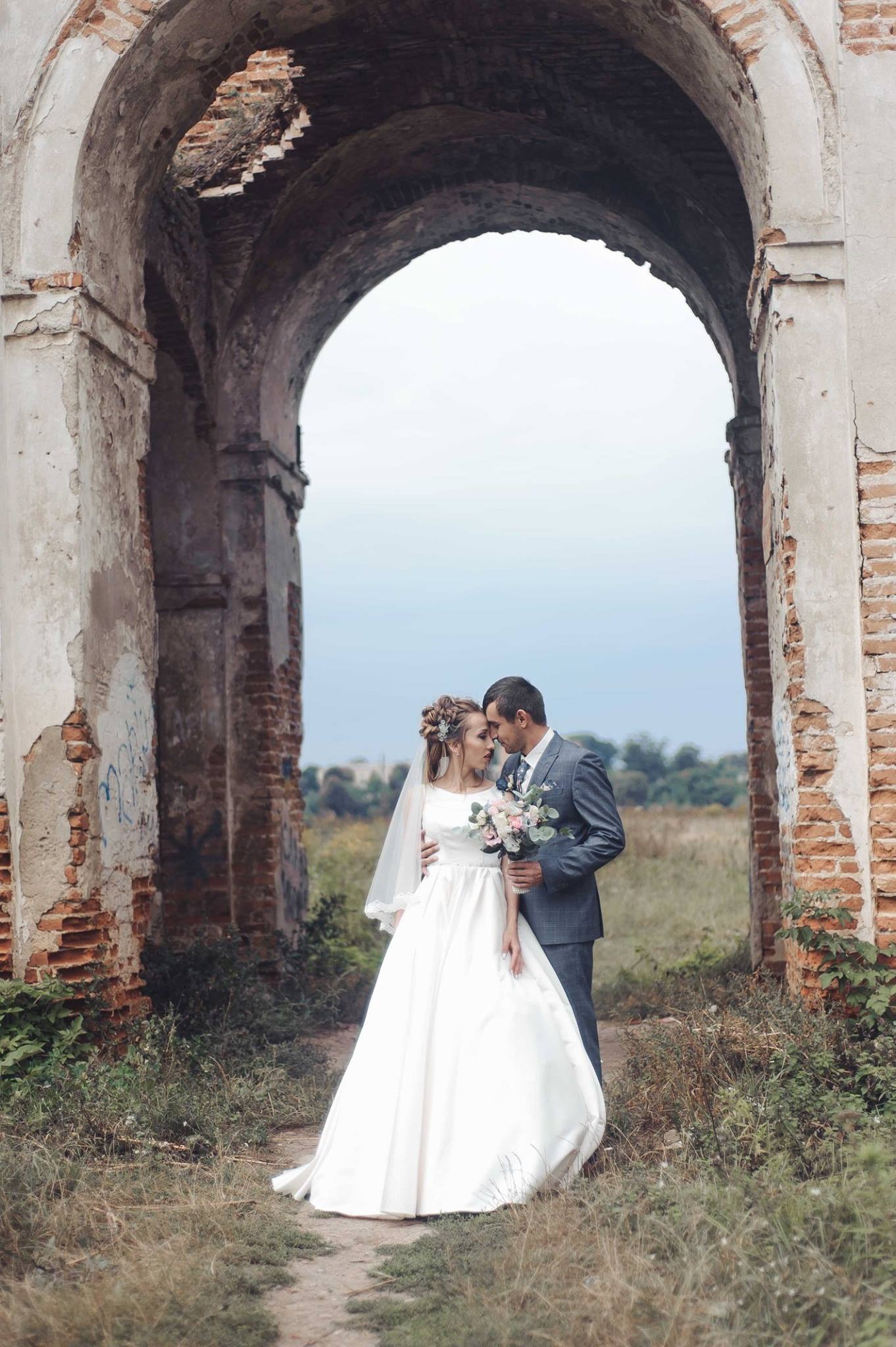 Para młoda w objęciach, stojąca pod zrujnowanym ceglanym łukiem, panna młoda w białej sukni z bukietem, pan młody w szarym garniturze.