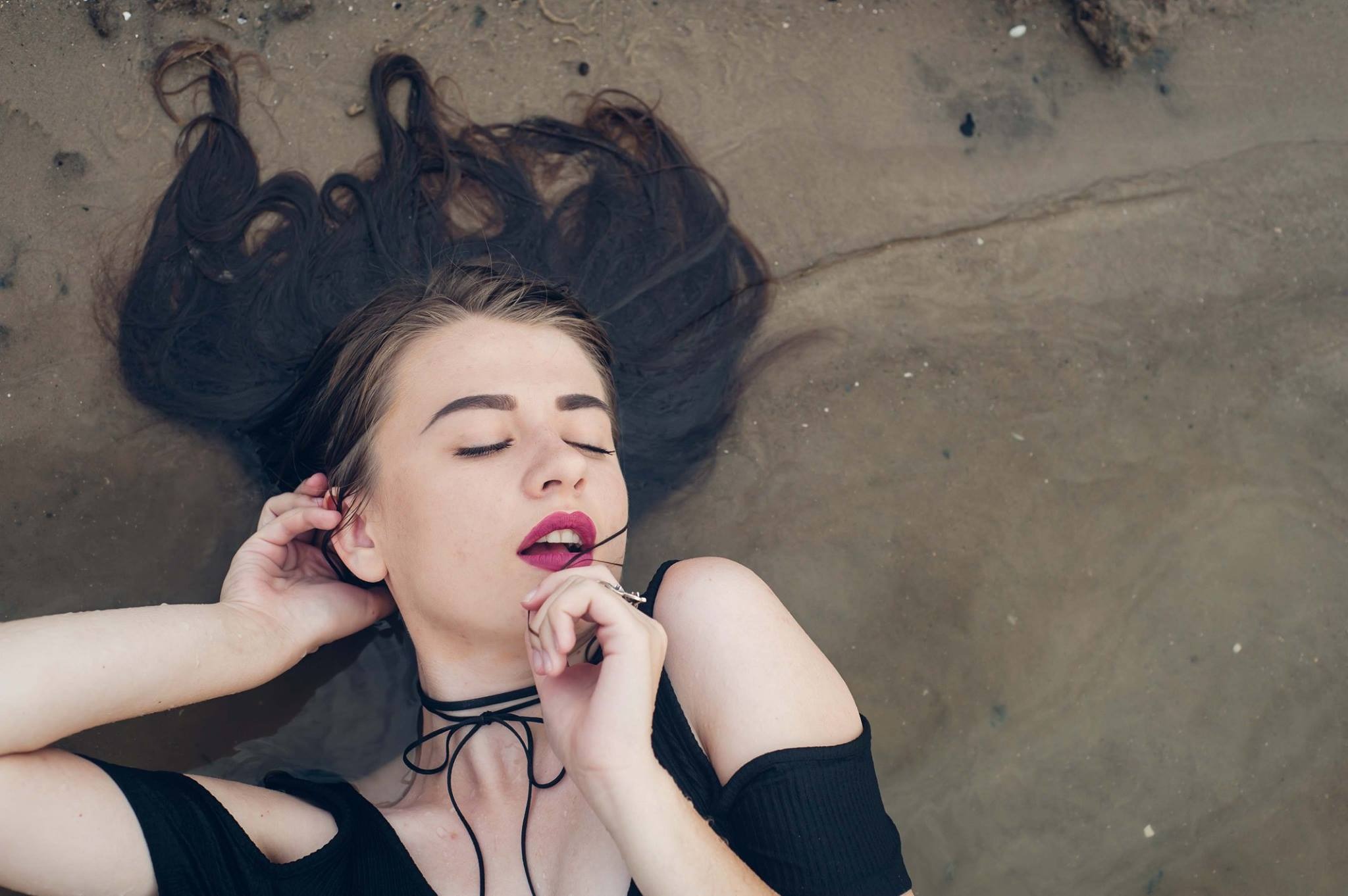 Portret kobiety leżącej w płytkiej wodzie, z rozpuszczonymi ciemnymi włosami na piasku, czarnym chokerem na szyi i ciemną szminką na ustach.