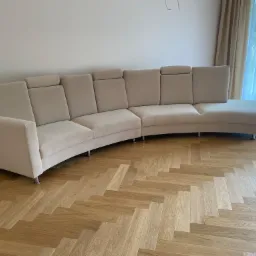 Beżowa, modułowa sofa z regulowanymi zagłówkami, ustawiona na parkiecie w jodełkę, z widokiem na okno z delikatnymi zasłonami.
