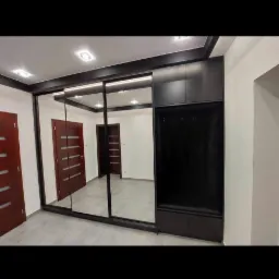 Czarna szafa wnękowa z przesuwnymi lustrzanymi drzwiami, wbudowana w przedpokoju z widocznymi drzwiami wejściowymi.