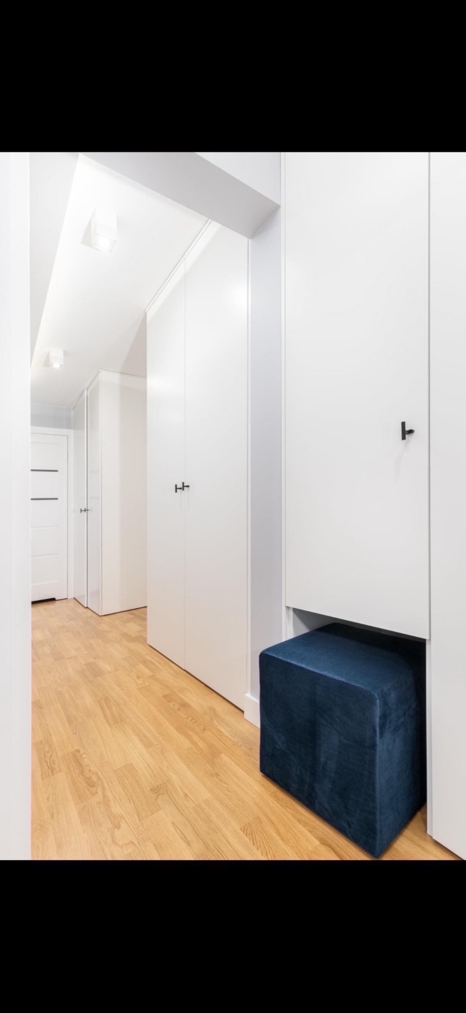 Biały korytarz z zabudowanymi szafami z minimalistycznymi, czarnymi uchwytami i drewnianą podłogą; ciemnoniebieski, welurowy puf wbudowany w jedną z szaf.