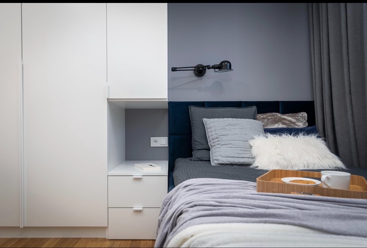 Nowoczesna sypialnia z zabudową meblową: biała szafa zintegrowana z szafką nocną, niebieskie tapicerowane wezgłowie, czarna lampka ścienna i drewniana taca z przekąskami na łóżku.