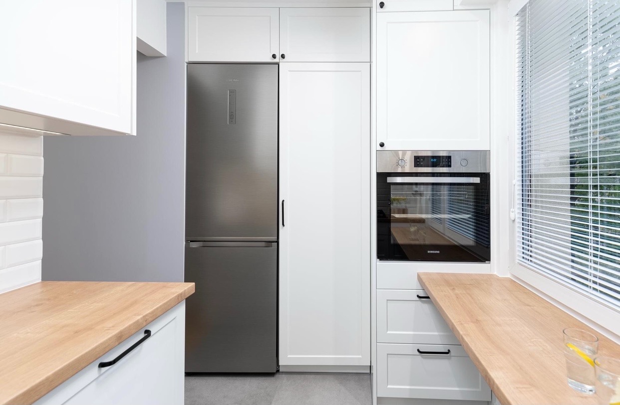 Nowoczesna, biała kuchnia z drewnianymi blatami, stalową lodówką Samsung i piekarnikiem w zabudowie, oświetlona naturalnym światłem zza żaluzji okiennych.