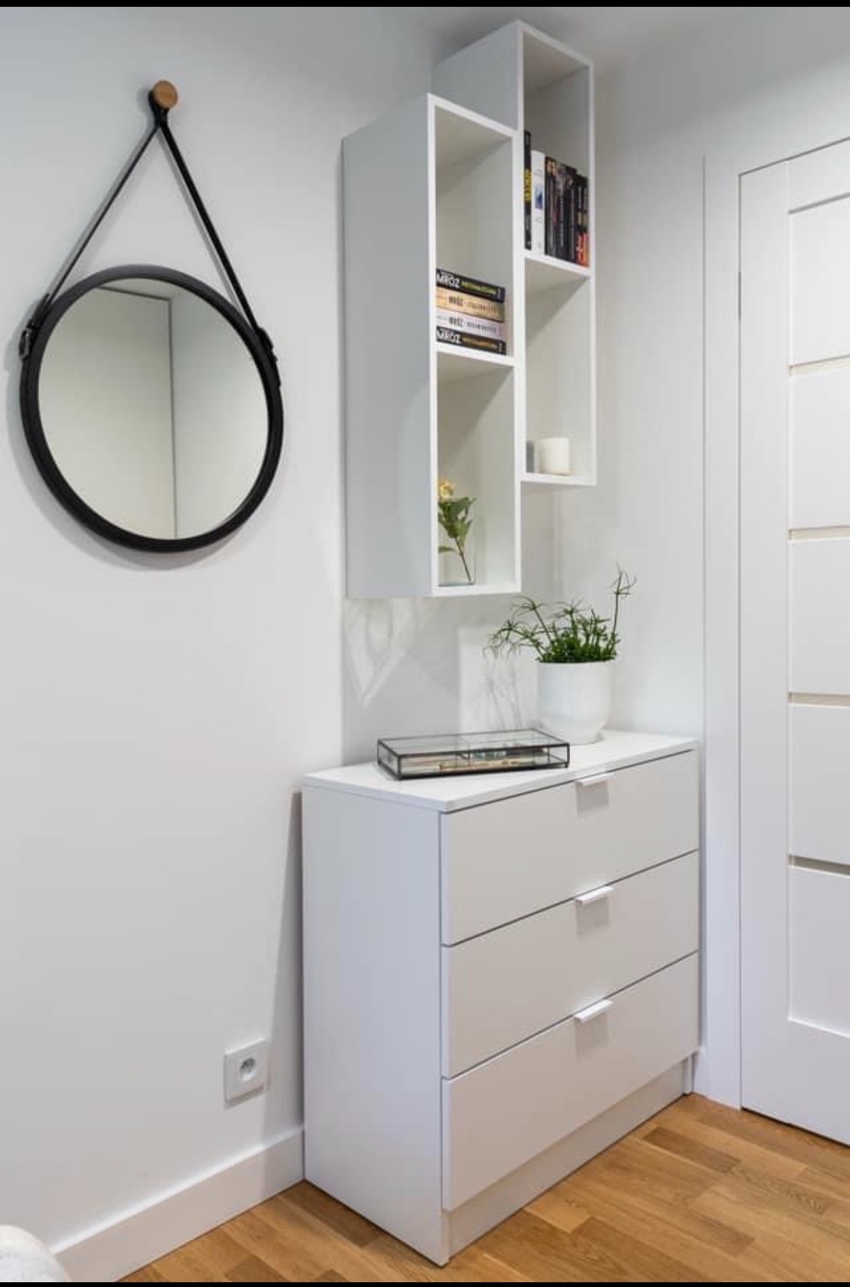 Biała komoda z trzema szufladami, na niej szklane pudełko i doniczka z rośliną, nad nią wisząca biała półka z książkami i okrągłe lustro w czarnej ramie na ścianie, obok białe drzwi.