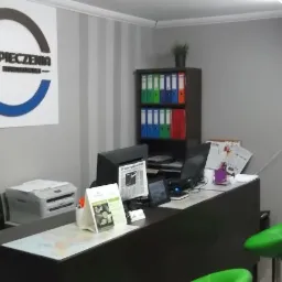 Biuro ubezpieczeniowe z logo firmy na ścianie, biurkiem z komputerem i zielonymi krzesłami barowymi.