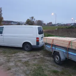 Biały samochód dostawczy z przyczepą wypełnioną drewnianymi belkami, zabezpieczonymi pasami, na tle zabudowy mieszkalnej i pochmurnego nieba.