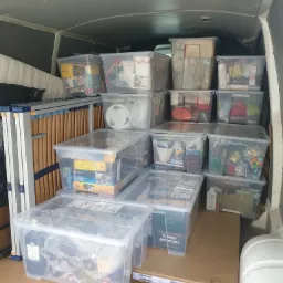 Wnętrze furgonetki wypełnionej przezroczystymi plastikowymi pojemnikami z różnymi przedmiotami, w tle widoczny złożony stelaż łóżka