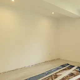 Pokój po malowaniu z nowo ułożonymi panelami podłogowymi, zabezpieczonymi kocem w rogu, widoczne gniazdka elektryczne i listwy przypodłogowe w trakcie montażu.