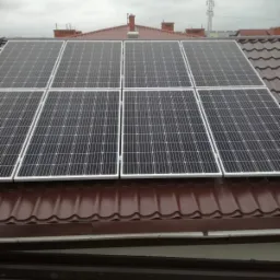 Panele słoneczne w białych ramach zamontowane na brązowym dachu z dachówki ceramicznej, w tle słup wysokiego napięcia i pochmurne niebo.