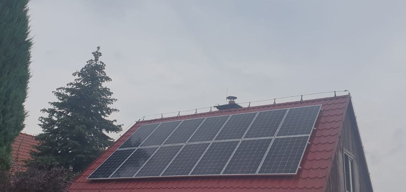 Panele słoneczne na dachu domu z czerwoną dachówką, widoczne drzewa iglaste i pochmurne niebo