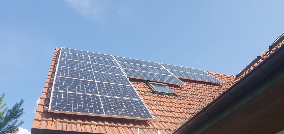 Panele słoneczne na dachu z czerwonej dachówki, umieszczone nad oknem dachowym, widoczne na tle błękitnego nieba.