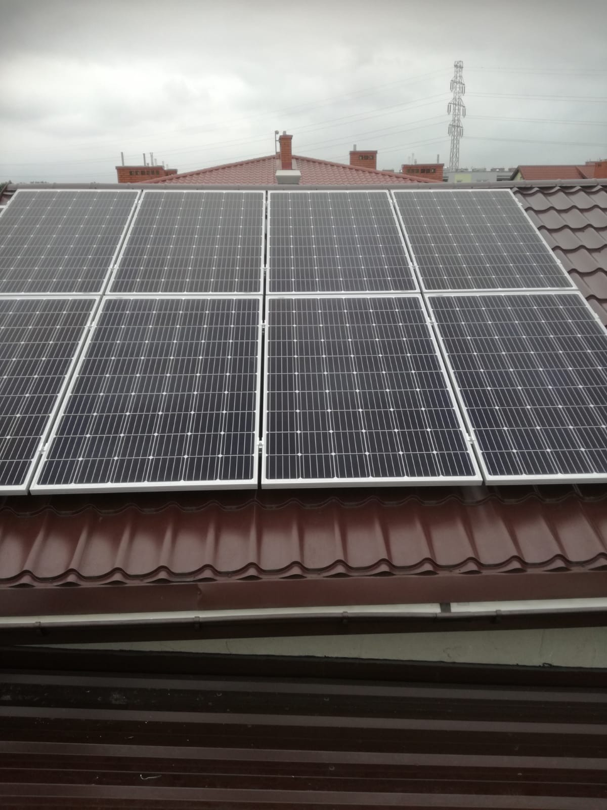 Panele słoneczne w białych ramach zamontowane na brązowym dachu z dachówki ceramicznej, w tle słup wysokiego napięcia i pochmurne niebo.
