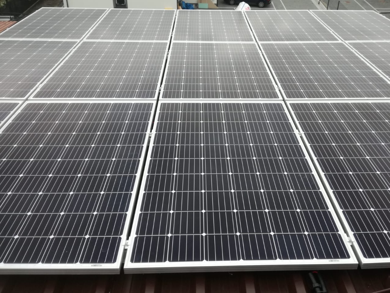 Rząd paneli słonecznych z białymi ramami, zainstalowanych na brązowym dachu z widocznymi przewodami w dolnym rogu, z fragmentem parkingu w tle.