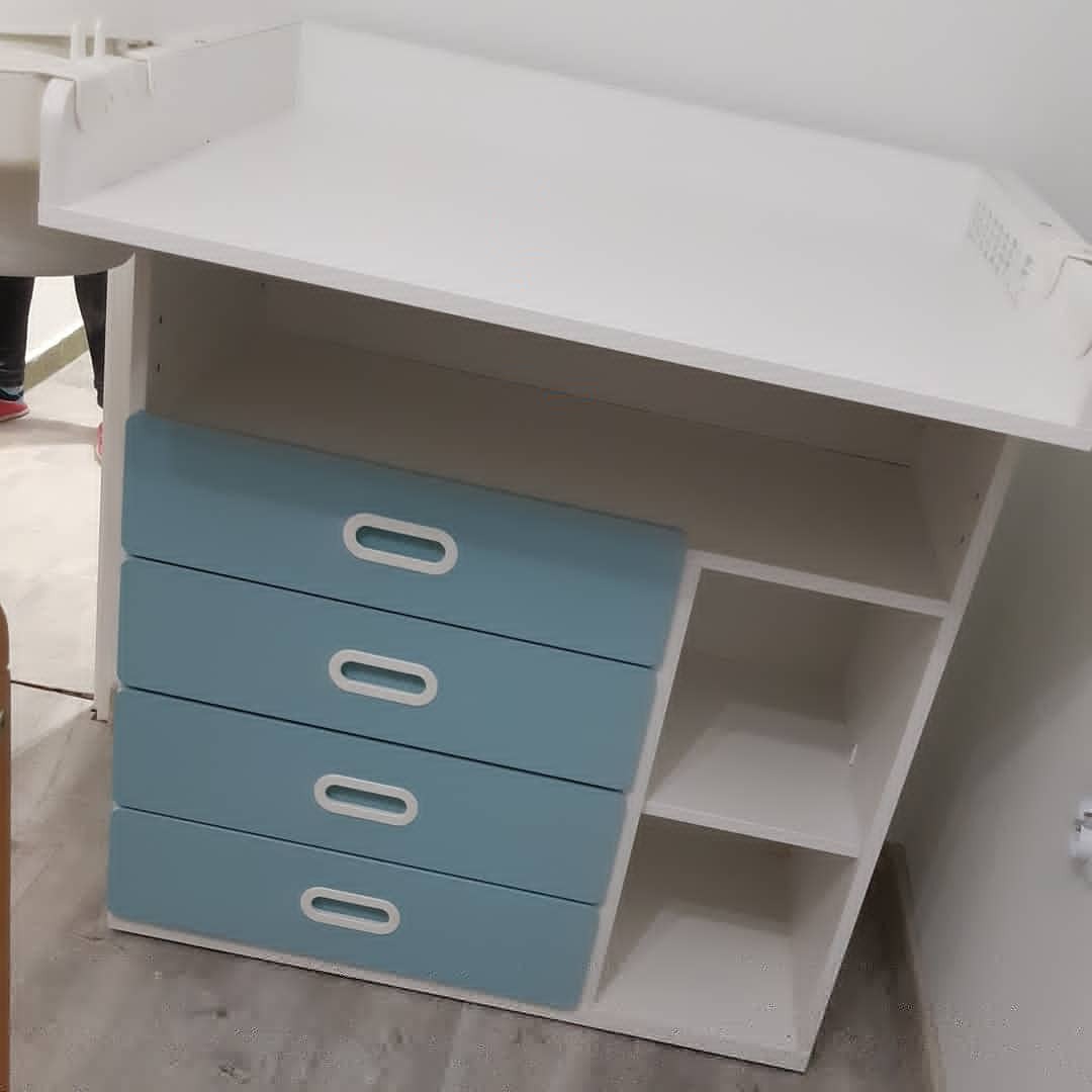 Biała komoda dziecięca z czterema błękitnymi szufladami z owalnymi uchwytami, widoczne półki po prawej stronie, ujęcie z dołu.