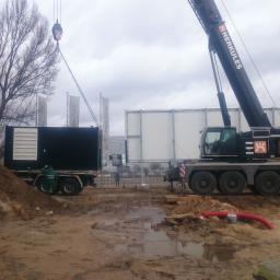 Electro-Power Sp. z o.o. - Mobilny generator prądu podnoszony przez dźwig Herkules na placu budowy, widoczny pracownik w zielonym kombinezonie, ziemia i kałuże.