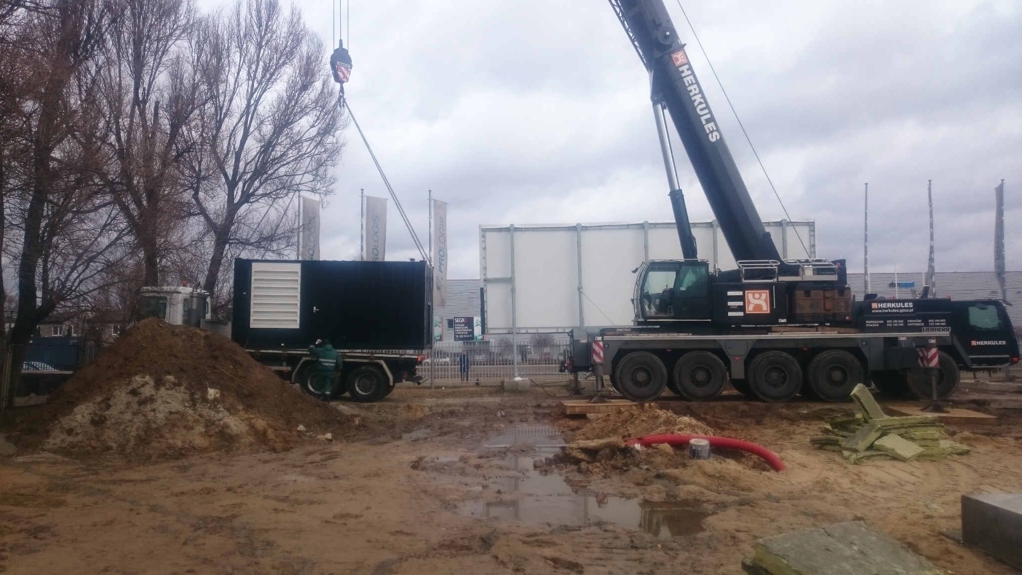 Mobilny generator prądu podnoszony przez dźwig Herkules na placu budowy, widoczny pracownik w zielonym kombinezonie, ziemia i kałuże.
