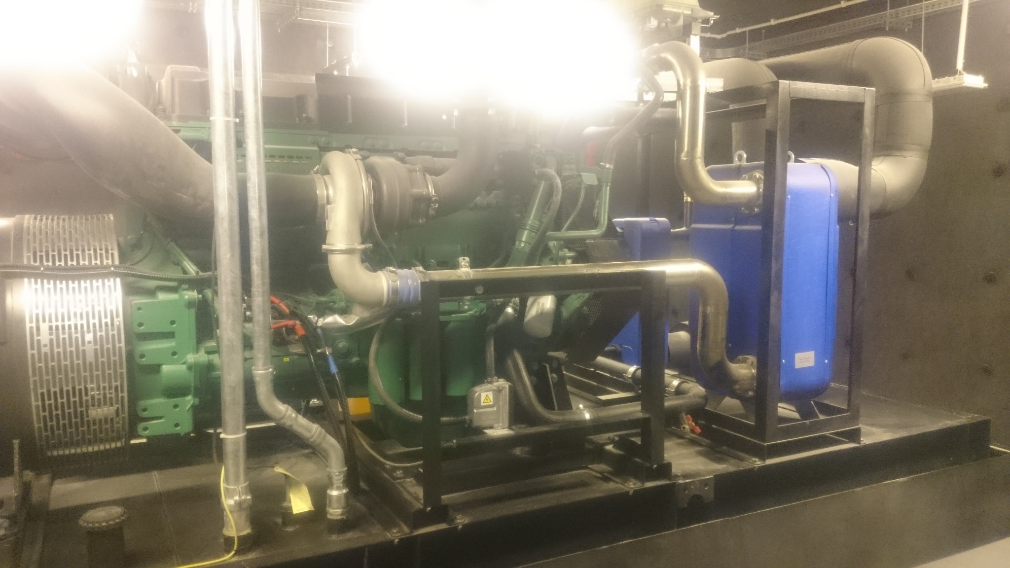 Zielony agregat prądotwórczy z metalową ramą i systemem rur w pomieszczeniu technicznym.