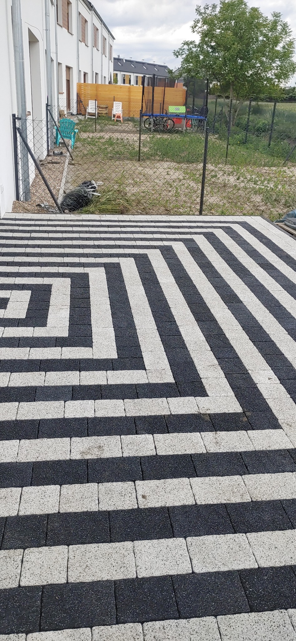 Ułożona kostka brukowa w czarno-białe pasy tworzące geometryczny wzór, widok z góry na tle trawnika, ogrodzenia i budynku mieszkalnego.