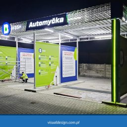 VISUAL Sp. z o.o. - Nowoczesna konstrukcja myjni samochodowej z logo 'B Wash' w Ostrowcu Świętokrzyskim, oświetlona ledami. Widoczny pracownik w odblaskowej kamizelce.