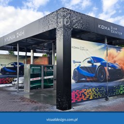 VISUAL Sp. z o.o. - Nowoczesna konstrukcja myjni samochodowej KOMA CAR CENTER z perforowanymi elementami i reklamami aut. Widoczny automat myjący i zaparkowany samochód.