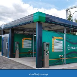 VISUAL Sp. z o.o. - Zadaszona, modułowa konstrukcja myjni samochodowej z panelami informacyjnymi i automatami płatniczymi. Widoczny samochód i zieleń w tle.