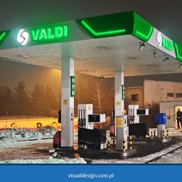 VISUAL Sp. z o.o. - Stacja paliw Valdi nocą, z zielonym neonowym oświetleniem. Widoczny budynek stacji, dystrybutory i częściowo ośnieżony teren. W tle budynki i drzewa.