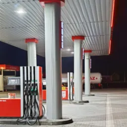 Nowoczesna stacja paliw nocą: białe kolumny z czerwonymi akcentami podświetlone ledami, dystrybutory paliwa i cysterna w tle. Minimalistyczny design.