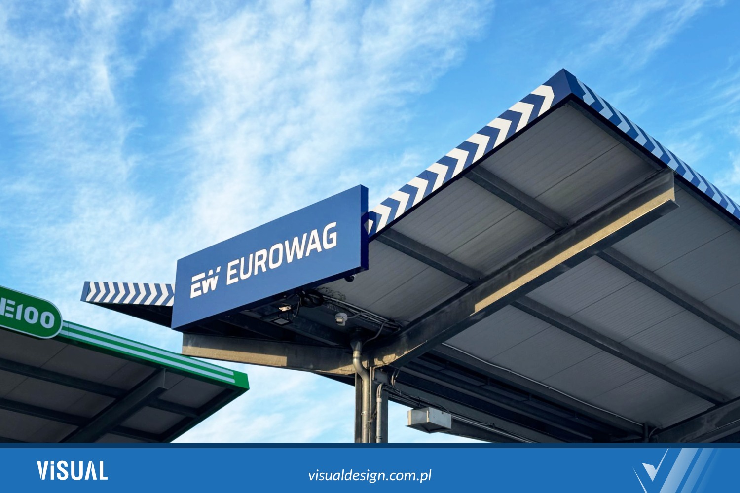 Stylowy pylon z logo EW EUROWAG na tle błękitnego nieba, z elementami konstrukcji stalowej i dekoracyjnymi pasami. Widoczne detale architektoniczne i branding.