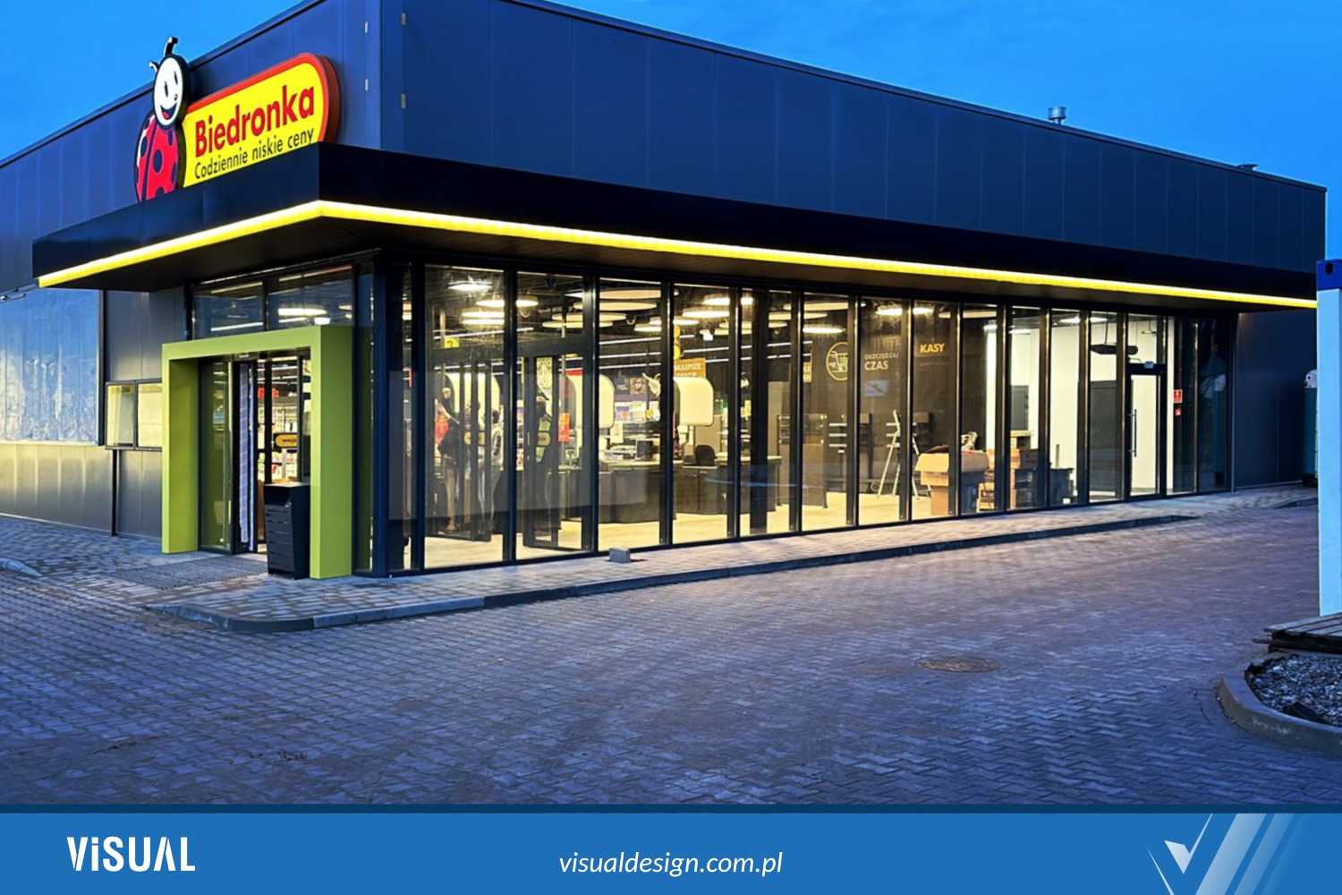 Elewacja nowoczesnego sklepu Biedronka z przeszkloną fasadą i charakterystycznym logo, oświetlona listwami LED, widoczna z zewnątrz na tle wieczornego nieba.