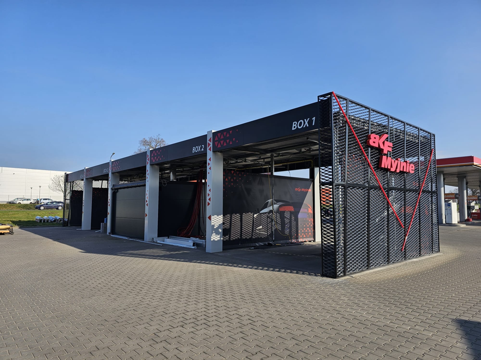 Nowoczesna, otwarta konstrukcja myjni samochodowej z czarnymi panelami i czerwonym akcentem kolorystycznym, oznaczona numerami stanowisk na tle błękitnego nieba.