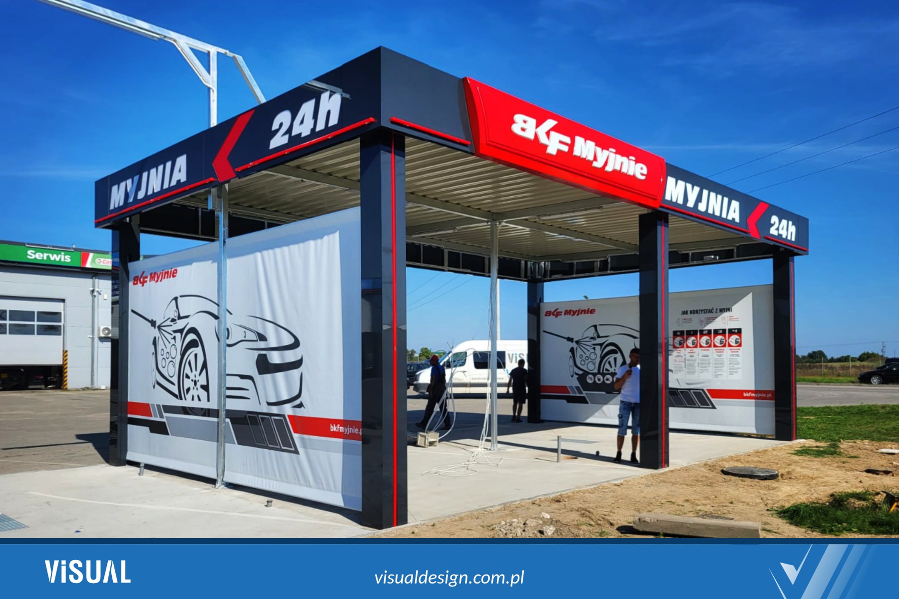 Zadaszona, samoobsługowa myjnia samochodowa z grafiką auta i logo 'BKF Myjnie', widoczna z zewnątrz w słoneczny dzień. Konstrukcja w kolorach szarym, czarnym i czerwonym.