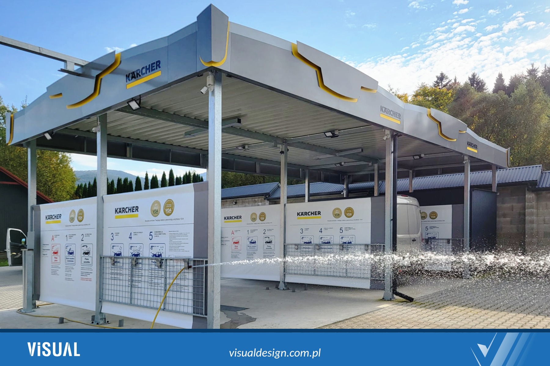 Nowoczesna, zadaszona myjnia samochodowa Kärcher z instrukcjami mycia na stanowiskach. Woda tryska z węża, co sugeruje działanie. Tło: drzewa, błękitne niebo.