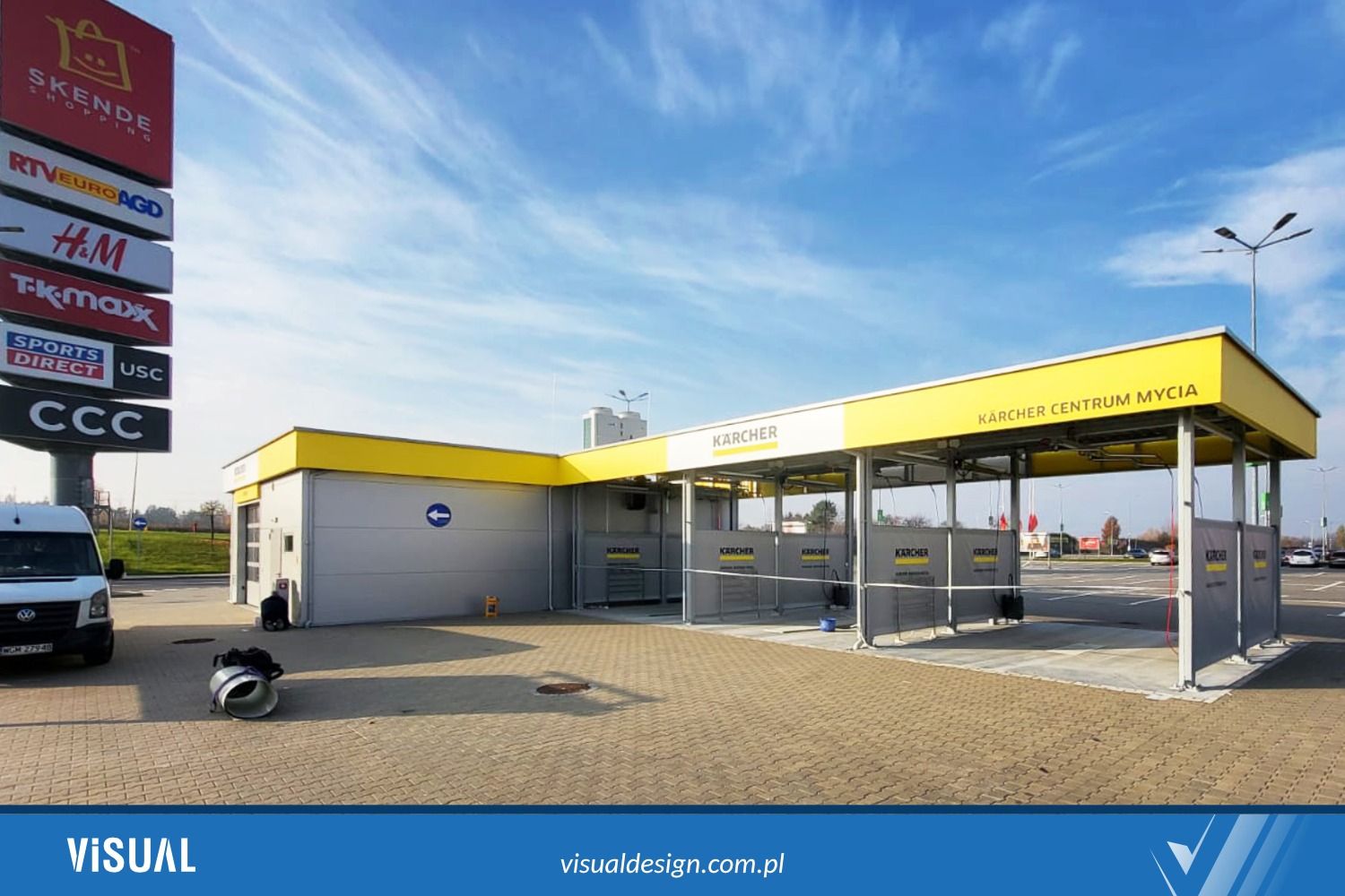 Centrum mycia Kärcher z żółto-szarą konstrukcją, widoczne logo i nazwa. Na pierwszym planie fragment parkingu z kostki brukowej, w tle budynki centrum handlowego.