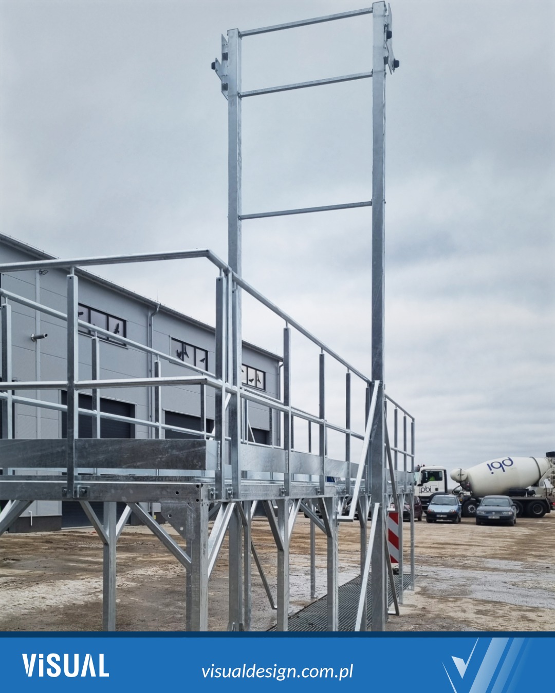 Metalowa konstrukcja schodów z platformą, ocynkowana, z barierkami, prowadząca do wejścia do hali przemysłowej. W tle zaparkowane pojazdy i pochmurne niebo.