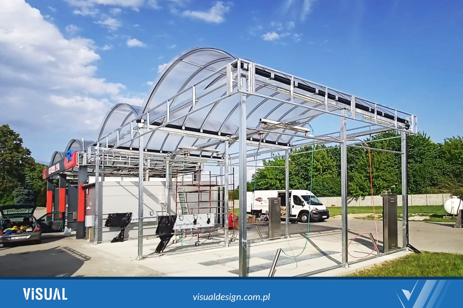 Konstrukcja stalowa myjni samochodowej z przezroczystym zadaszeniem, widok z zewnątrz, w tle zaparkowany biały bus i zieleń drzew.
