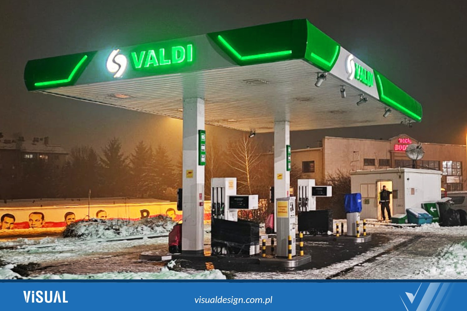 Stacja paliw Valdi nocą, z zielonym neonowym oświetleniem. Widoczny budynek stacji, dystrybutory i częściowo ośnieżony teren. W tle budynki i drzewa.
