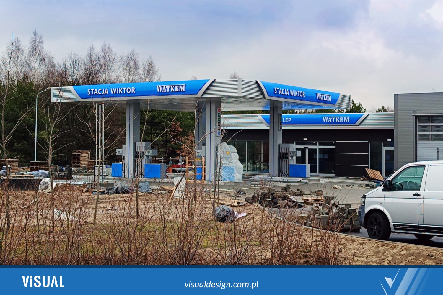 Stacja paliw 'Stacja Wiktor WATKEM' z dystrybutorami, w trakcie prac wykończeniowych terenu, biały bus z boku, w tle drzewa. Błękitno-srebrna kolorystyka.