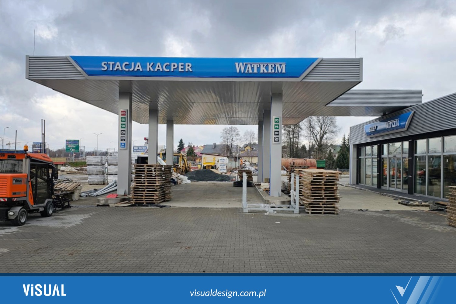 Stacja benzynowa w trakcie prac budowlanych: widoczne palety, maszyny budowlane, logo 'Stacja Kacper' i 'Watkem' na zadaszeniu. Szaro-błękitna kolorystyka.