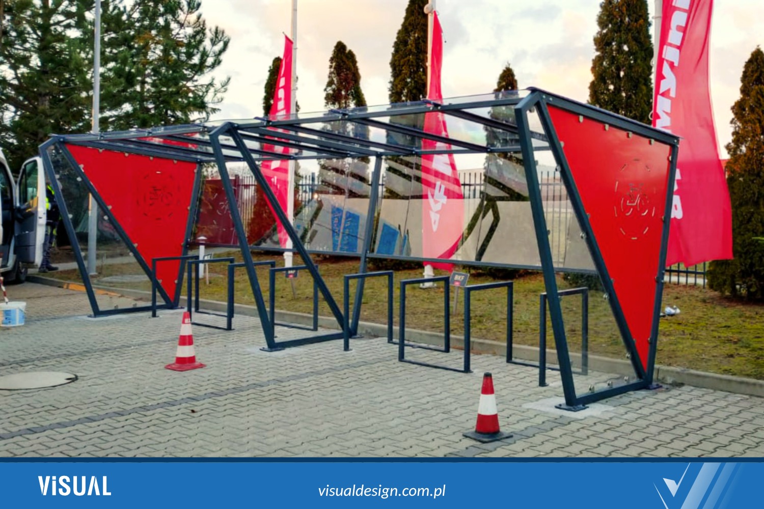 Zadaszony, nowoczesny parking dla rowerów z czerwonymi, trójkątnymi panelami bocznymi i przezroczystym dachem, ustawiony na kostce brukowej. Widoczne flagi w tle.